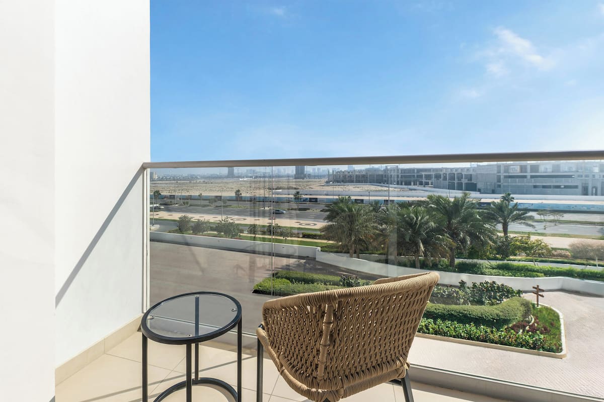 A balcony with a wicker chair and a small table, overlooking a landscaped area with palm trees and a distant cityscape.