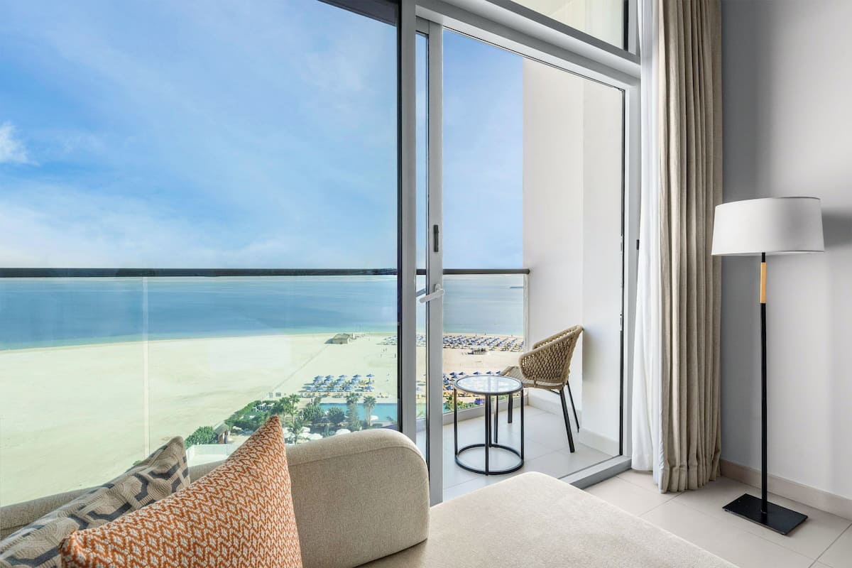 A balcony with a view of the beach, a sofa, a table, and a lamp.