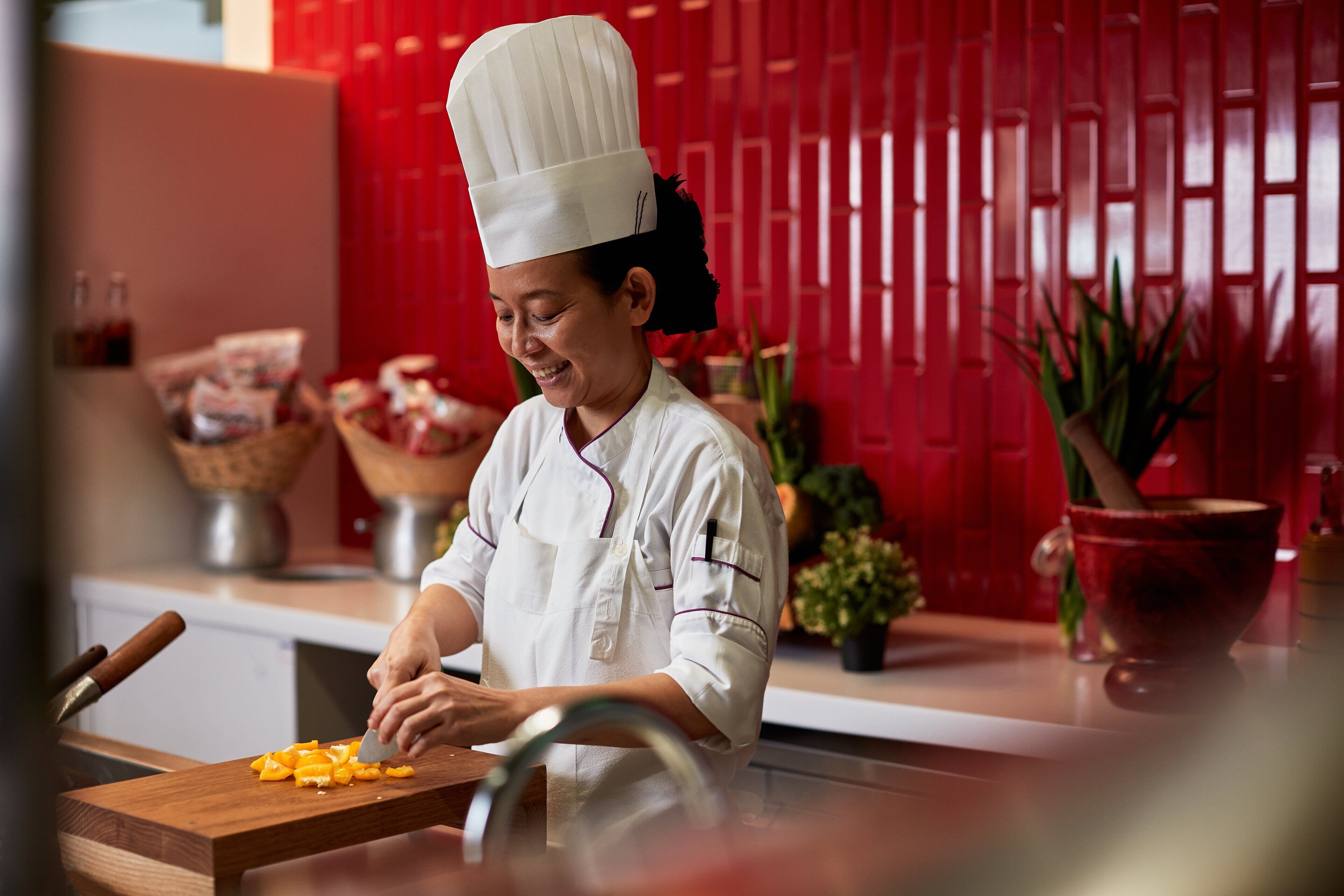 A chef in a white uniform with a tall hat, chopping vegetables on a wooden cutting board in a kitchen with red walls and various kitchen items in the background.