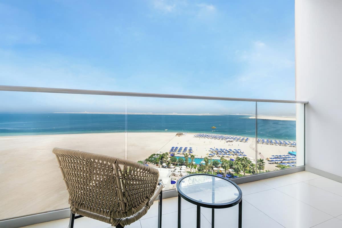 A balcony with a wicker chair and a small table, overlooking a beach with sun loungers and a pool.