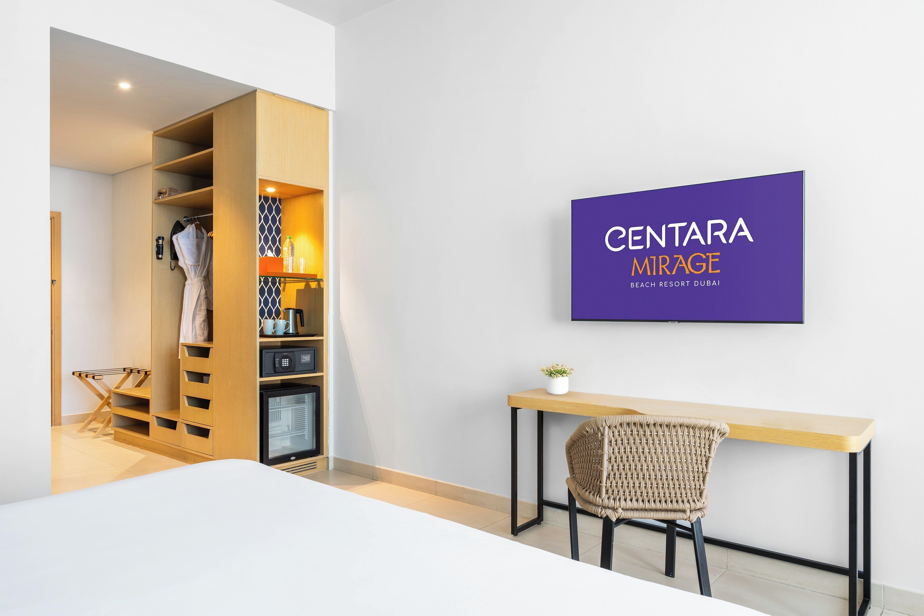 Une chambre d’hôtel moderne avec un bureau en bois, une chaise en osier, une armoire et un téléviseur fixé au mur qui affiche « CENTARA MIRAGE BEACH RESORT DUBAI ».