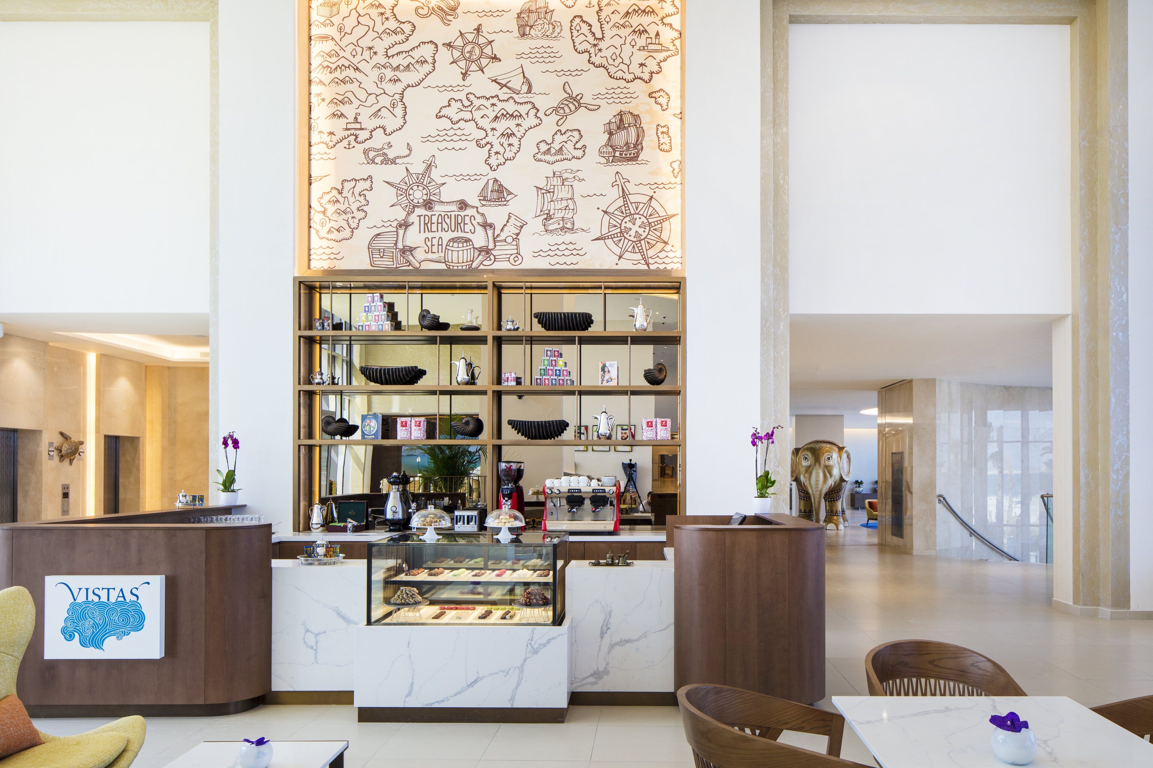 A modern hotel lobby with a reception desk, a coffee bar, and a display case of pastries.
