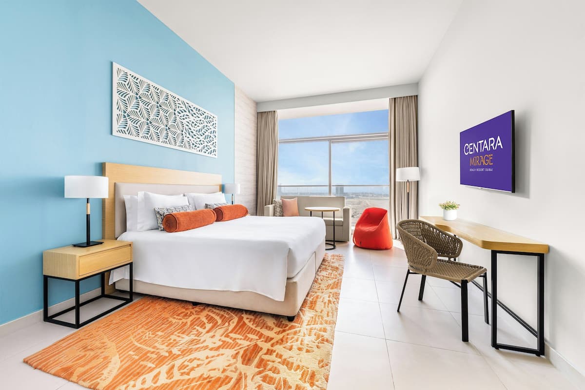A modern hotel room with a bed, a desk, a chair, a nightstand, a lamp, and a view of the sky.
