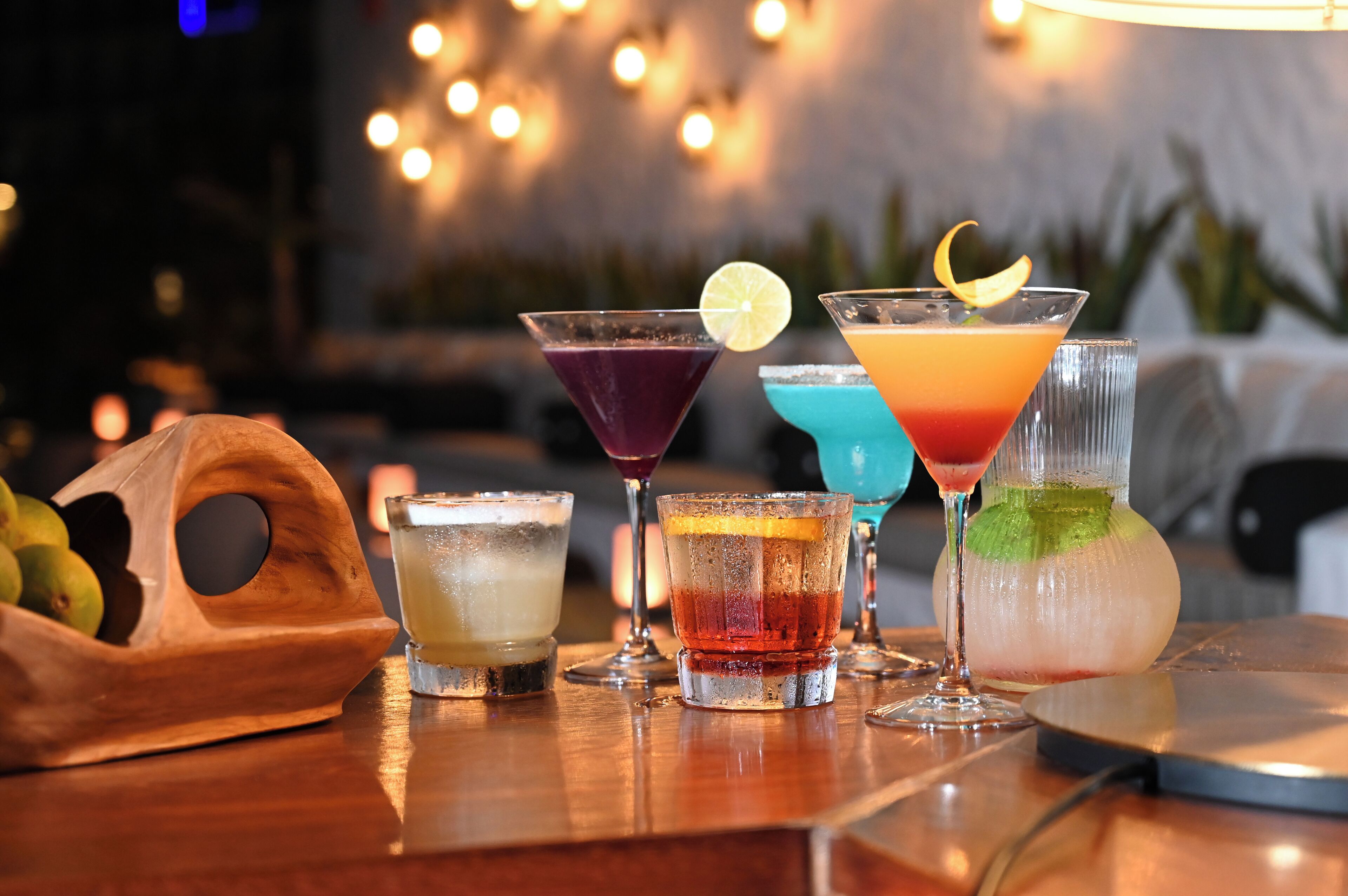 Una zona de bar con varios cócteles en vasos, un cuenco de frutas de madera y un fondo difuminado con luces.