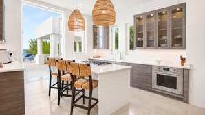 A modern kitchen with a central island, pendant lights, and a view to the outdoors. - Sand Villa – Greek-Inspired Elegance on the Shores of Long Bay, Anguilla (Long Bay Village)