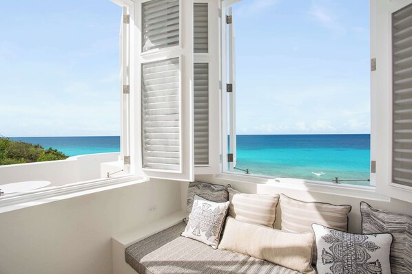 Un balcón con vistas al océano, una ventana con contraventanas blancas y una zona de estar con cojines estampados.