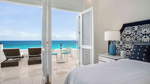 Une chambre avec un grand lit, une vue sur l’océan, un balcon avec des chaises et une table sur laquelle se trouvent une bouteille et des verres.