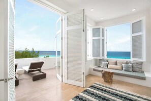 A balcony with a view of the sea, a seating area with cushions, and a large window with shutters.