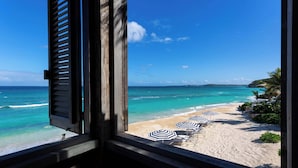 Une vue sur la plage depuis une fenêtre ornée de volets en bois, une plage de sable et des parasols de plage à rayures.