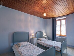 A room with two beds, a wooden ceiling, a window with curtains, and a radiator.