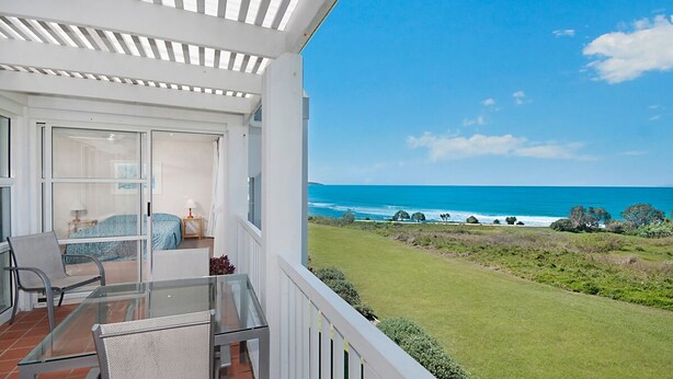 A balcony with a glass table and chairs, overlooking a grassy area and the ocean. - Quarterdeck 20 - Spectacular Ocean Views (Lennox Head)