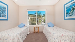 A bedroom with two single beds, a small wooden stool, and a window with a view of greenery and buildings. - Quarterdeck 20 - Spectacular Ocean Views (Lennox Head)