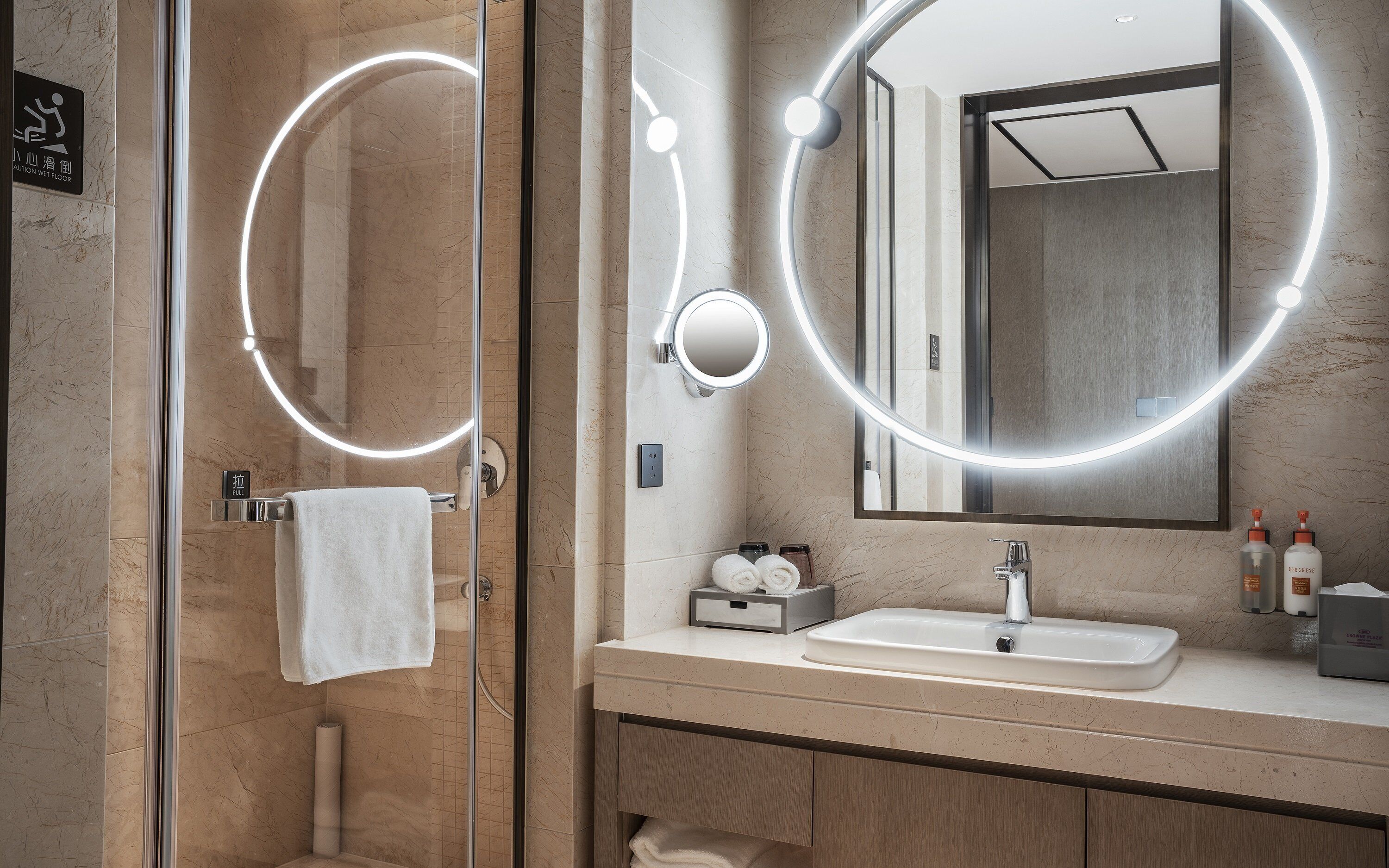 Un baño moderno con cabina de ducha de vidrio, tocador con lavabo y un espejo grande con iluminación circular.