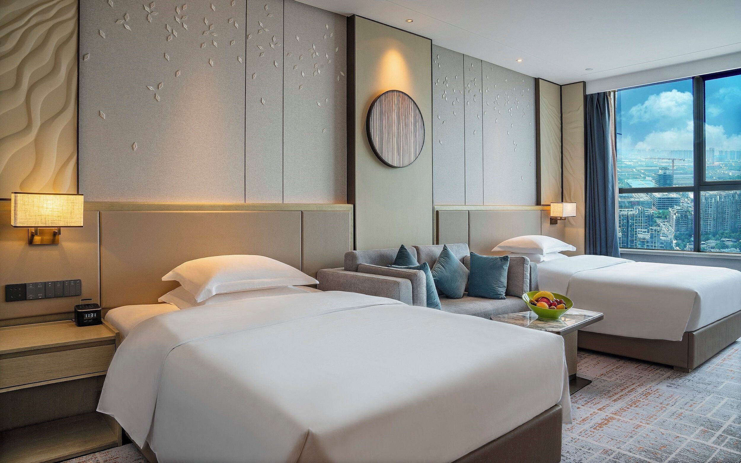 Une chambre d’hôtel avec deux lits, un canapé, une table d’appoint avec un panier de fruits, et une vue sur la ville à travers de grandes fenêtres.