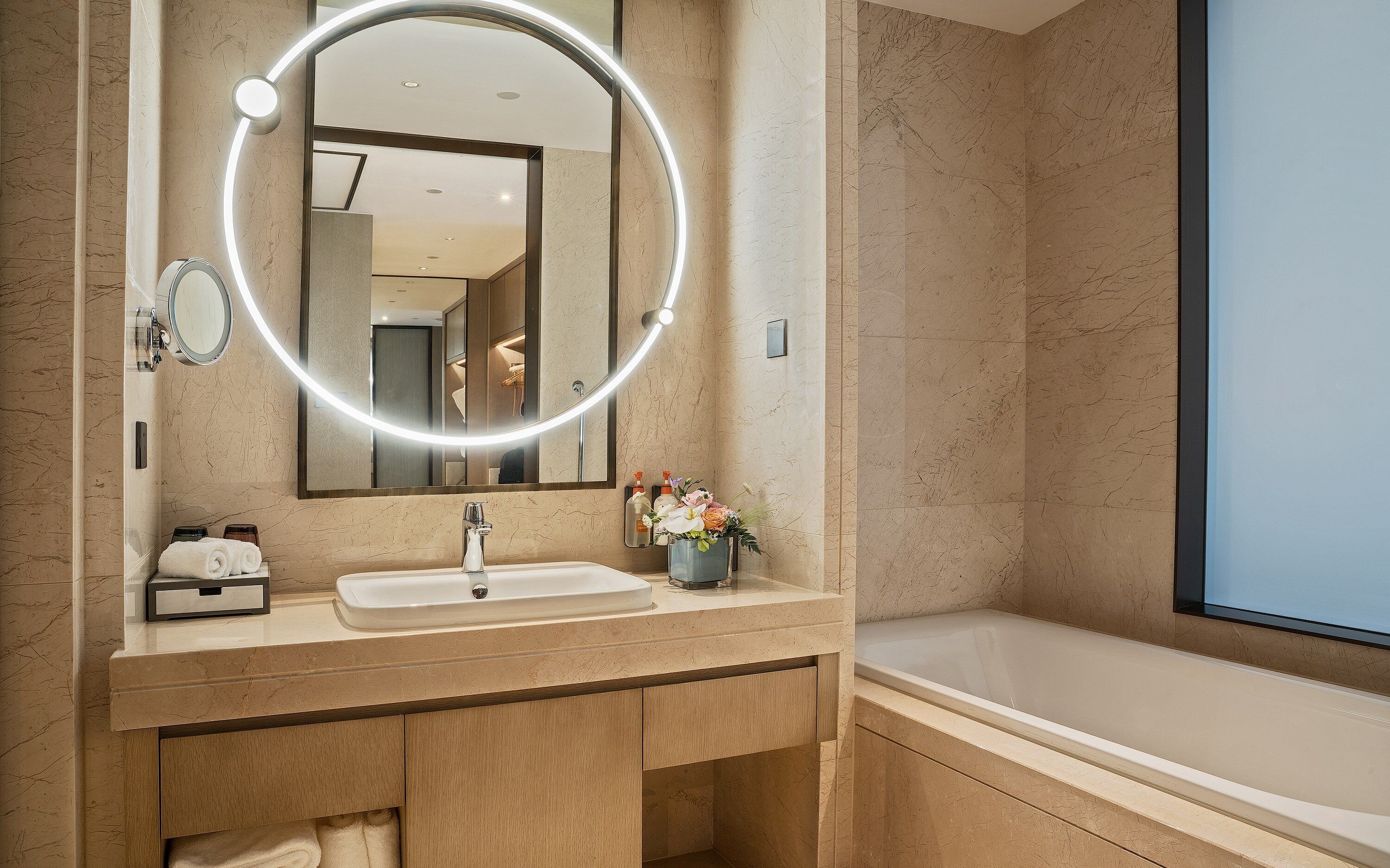 A modern bathroom with a marble vanity, a large mirror with lights, a bathtub, and a small flower arrangement.