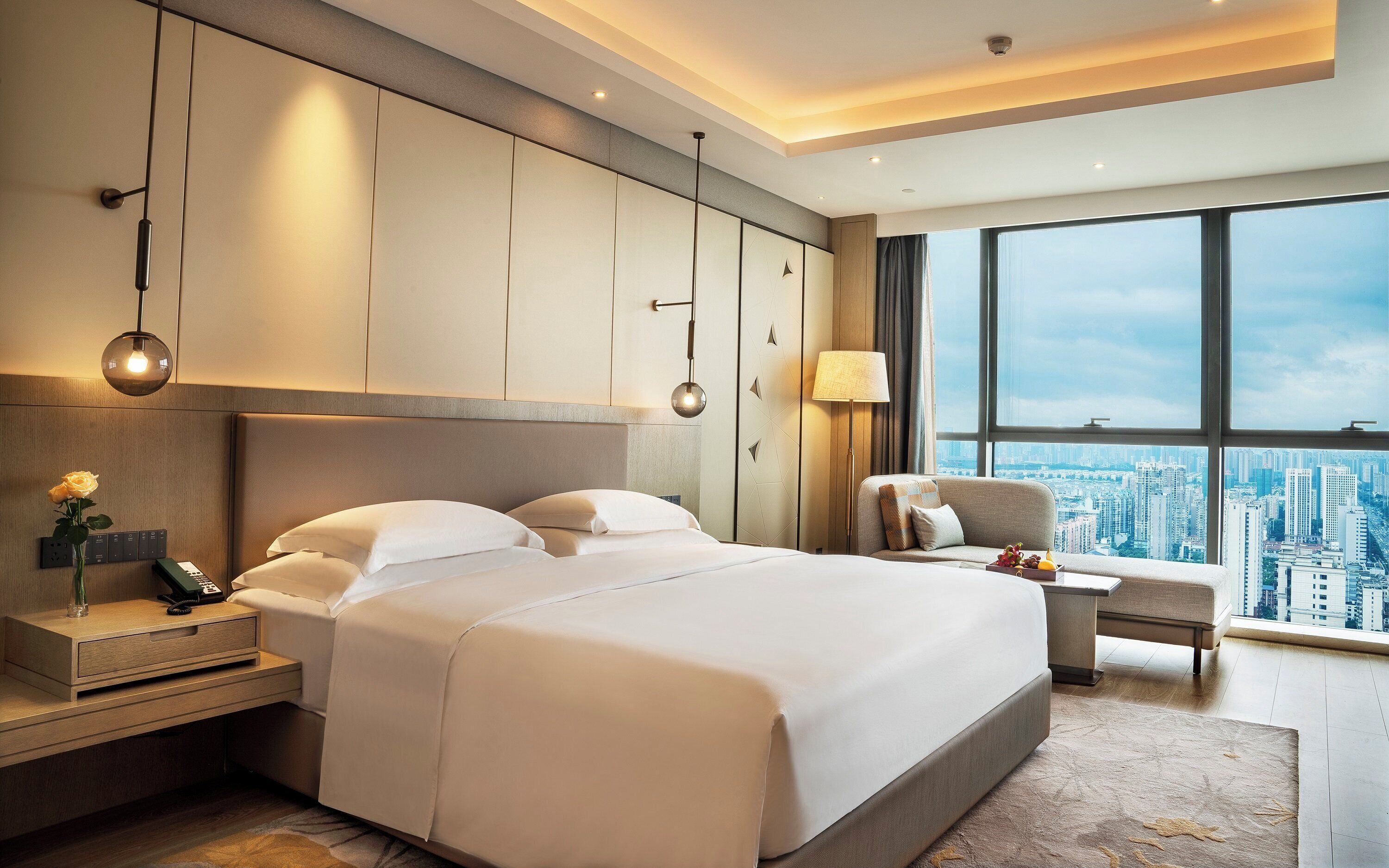 Quarto de hotel com cama grande, mesas de cabeceira, escrivaninha, cadeira e vista para a cidade.