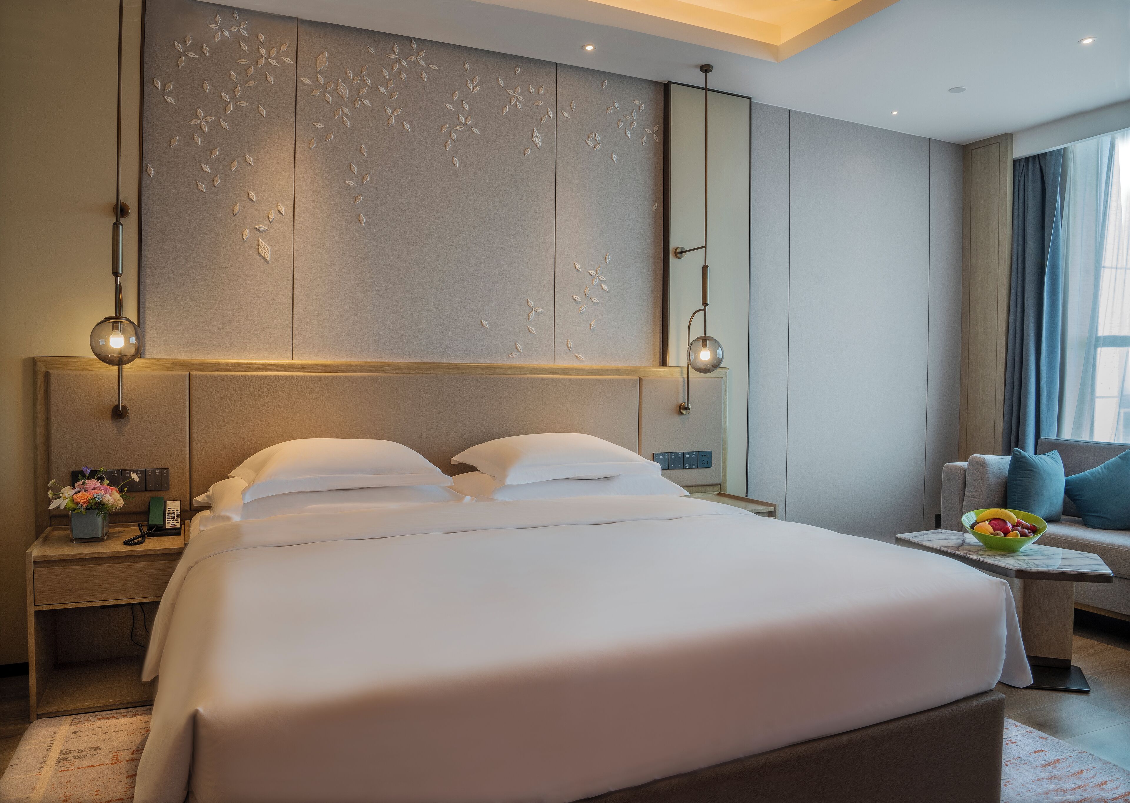 A modern hotel room with a large bed, bedside tables, a small seating area, and a decorative wall panel.