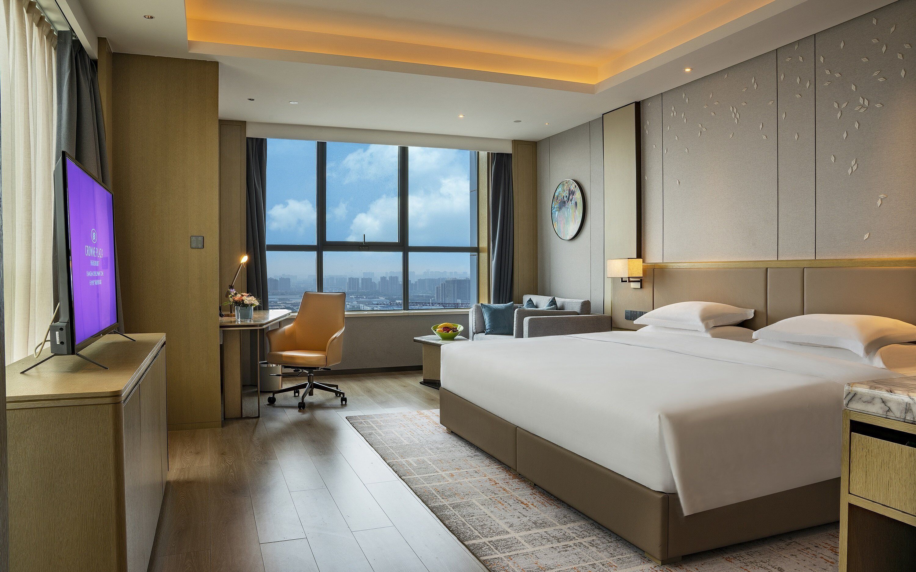 A hotel room with a large bed, a desk with a computer, a chair, and a view of the city through large windows.