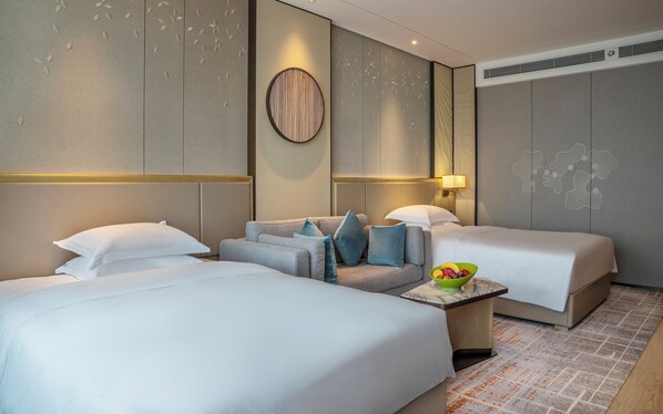 Une chambre d’hôtel avec deux lits, un canapé, une petite table sur laquelle se trouve un saladier rempli de fruits et un panneau décoratif au mur.