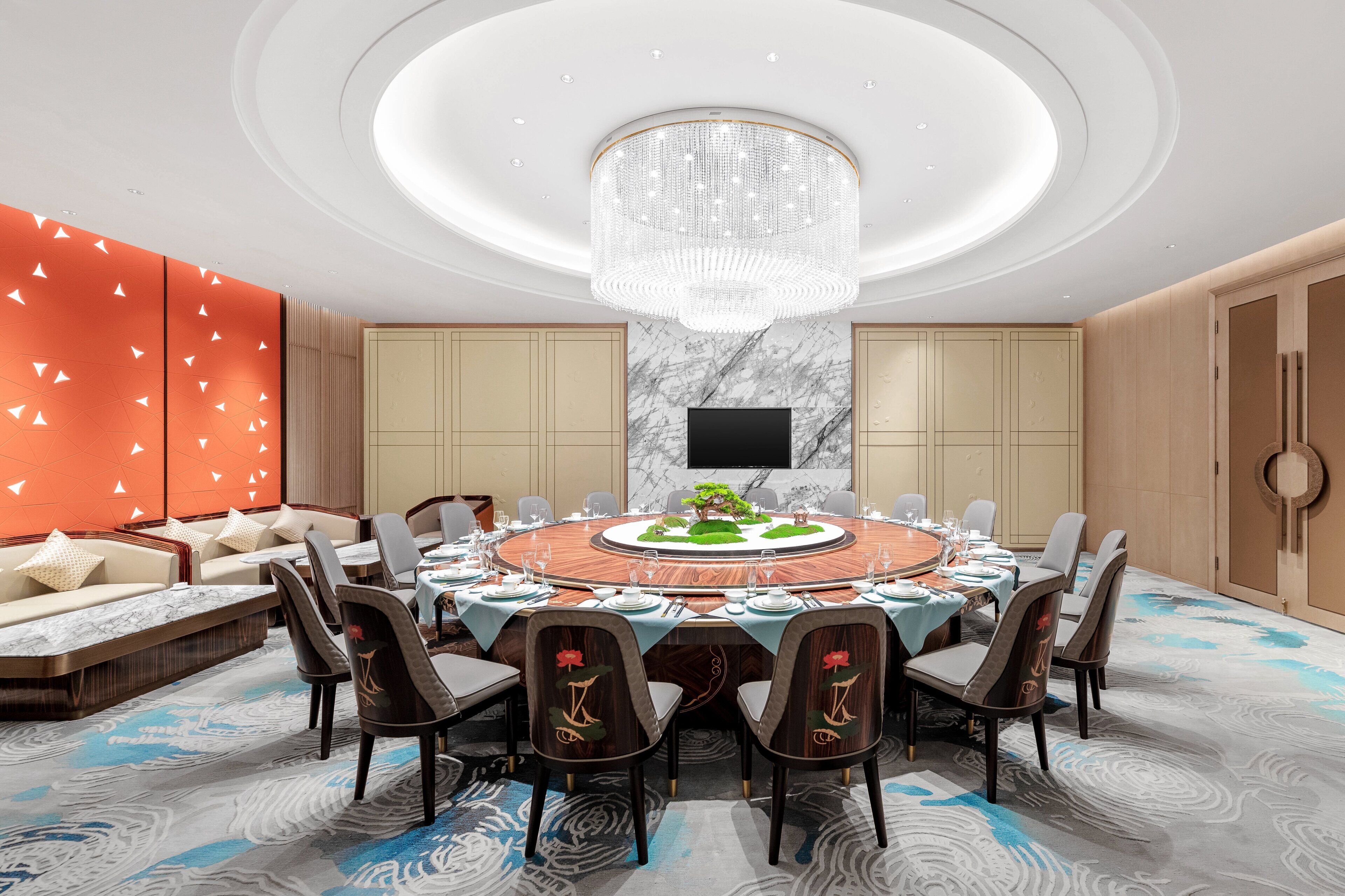 A modern conference room with a round table, comfortable seating, a large chandelier, and a flat-screen TV.