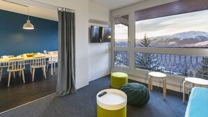 Une chambre d’hôtel moderne dotée d’un coin repas, d’une grande fenêtre offrant une vue sur les montagnes et d’un espace salon confortable avec des poufs.