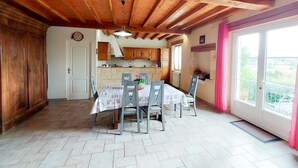 A dining area with a wooden ceiling, a kitchen in the background, and a view through a window.