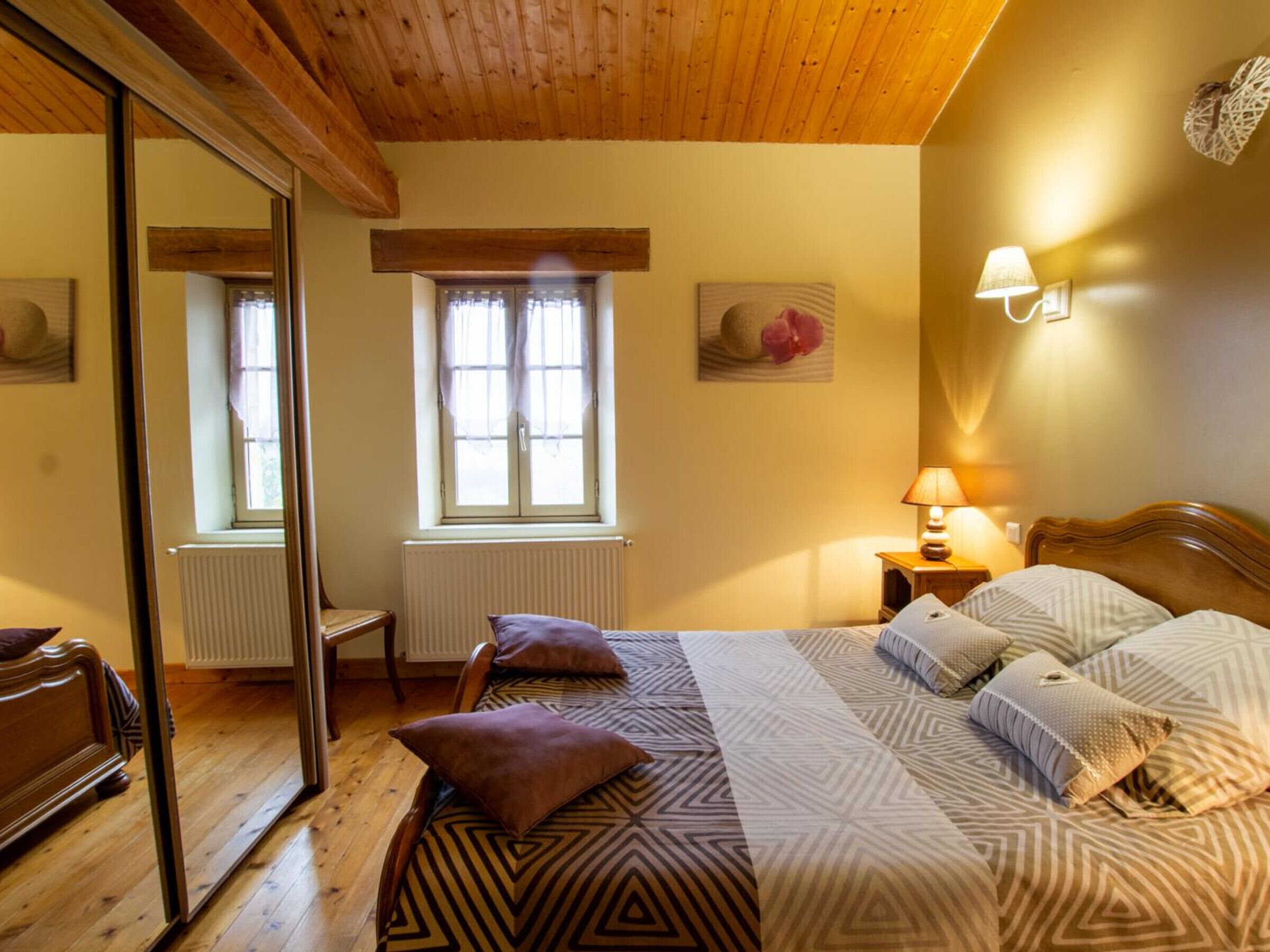 Ein Schlafzimmer mit Holzboden, einem Bett mit gemusterten Betttextilien, einem Nachttisch mit Lampe, einem Stuhl und einem Fenster mit Vorhängen.