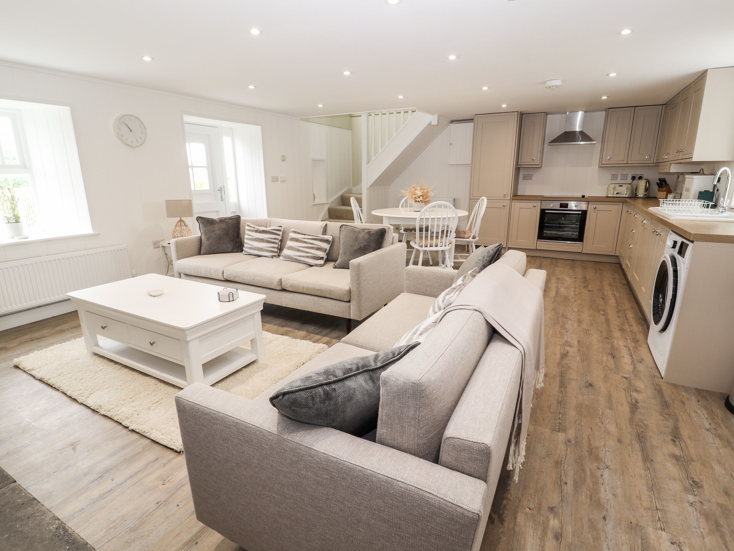 A modern living room with a sofa, coffee table, and a dining area with chairs. There is a kitchen with a stove, oven, and washing machine.