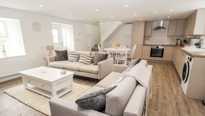 A modern living room with a sofa, coffee table, and a dining area with chairs. There is a kitchen with a stove, oven, and washing machine.