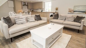 A modern living room with two beige sofas, a white coffee table, and a kitchen in the background.