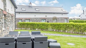 A stone building with a white facade and a grey roof, featuring a small garden with a hedge and outdoor seating area with wicker furniture. - Sgubor (Holyhead)