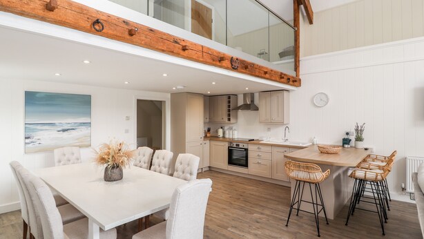 A modern kitchen with wooden beams, a dining area with white chairs, and a view of the sea through a large window. - Sgubor (Holyhead)