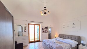 Una camera da letto con un letto, un comodino, una cassettiera, uno specchio, un lampadario e una porta scorrevole in vetro che dà su un balcone.