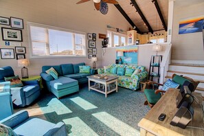 A cozy living room with blue furniture, a coffee table, and a TV. There are framed pictures on the wall and a window with blinds. - Oceanfront delight awaits at Livin on the Edge (Topsail Beach)