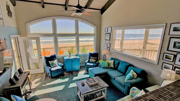Ein geräumiges Wohnzimmer mit großem Fenster zum Strand, einem Deckenventilator und verschiedenen Sitzgelegenheiten, darunter eine Couch und Sessel.
