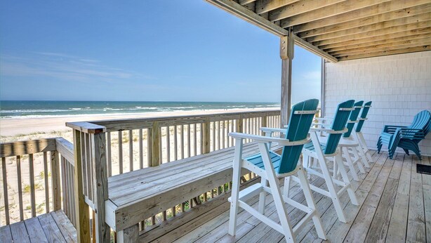 Terraza de madera con sillas Adirondack blancas y turquesa con vistas a una playa.