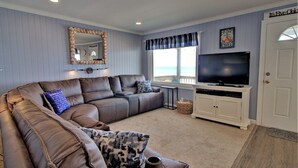 Una sala de estar con un sofá seccional, un televisor sobre un mueble blanco, un espejo grande y una ventana con vista al océano.