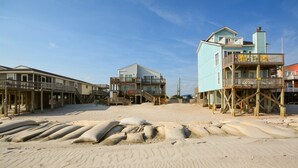 A beachfront property with multiple buildings, including a two-story house with a balcony and a larger building with a wooden deck. Sandbags are placed in the foreground, and the beach is visible in the background. - Oceanfront bliss awaits you at Cool Story, a 3BR retreat in North Topsail Beach. (North Topsail Beach)