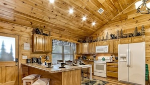 A wooden cabin interior with a kitchen area featuring wooden cabinets, a central island with stools, a microwave, a refrigerator, and a window with blinds.