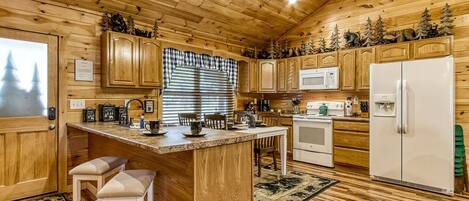 A wooden cabin interior with a kitchen area featuring wooden cabinets, a central island with stools, a microwave, a refrigerator, and a window with blinds.