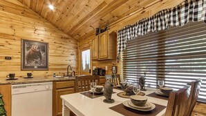 A wooden cabin interior with a dining table set for four, a kitchen area, and a framed bear picture on the wall.