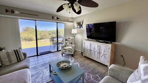 Una sala de estar con ventilador de techo, televisor, sofá, mesa de centro y vistas al exterior.