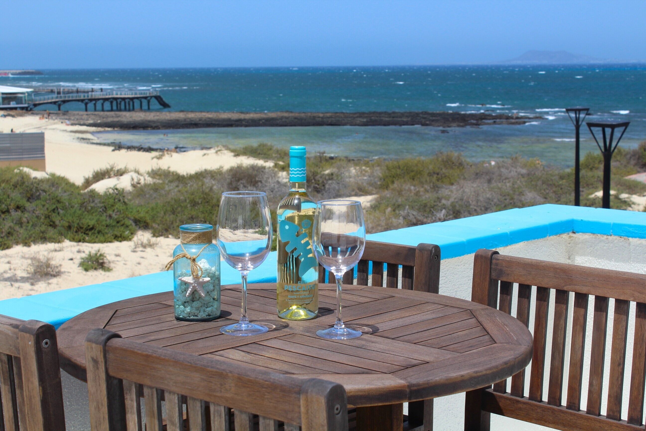 Een houten tafel met twee wijnglazen en een fles, met uitzicht op een strand en een pier op de achtergrond.