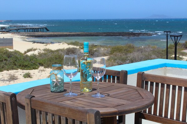 A wooden table with two wine glasses and a bottle, set against a beach view with a pier in the background. - Privilege Bay- Villa Eesha-sea view and natural park view. Beach front location. (La Oliva)