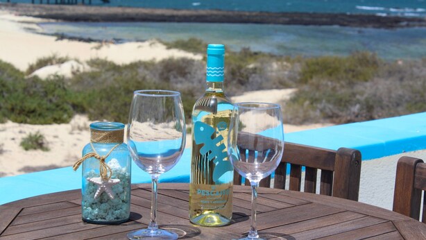 Ein Tisch mit einer Weinflasche, zwei Gläsern und einem Glas mit einem Seestern. Im Hintergrund sind Strand und Meer zu sehen.