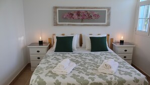 Una cama bien tendida con ropa de cama verde y blanca, dos mesitas de noche con velas, y una obra de arte floral enmarcada sobre la cama.