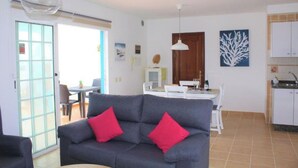 Una sala de estar con sofá gris, cojines rojos, una mesa de comedor y una zona de cocina con armarios blancos.