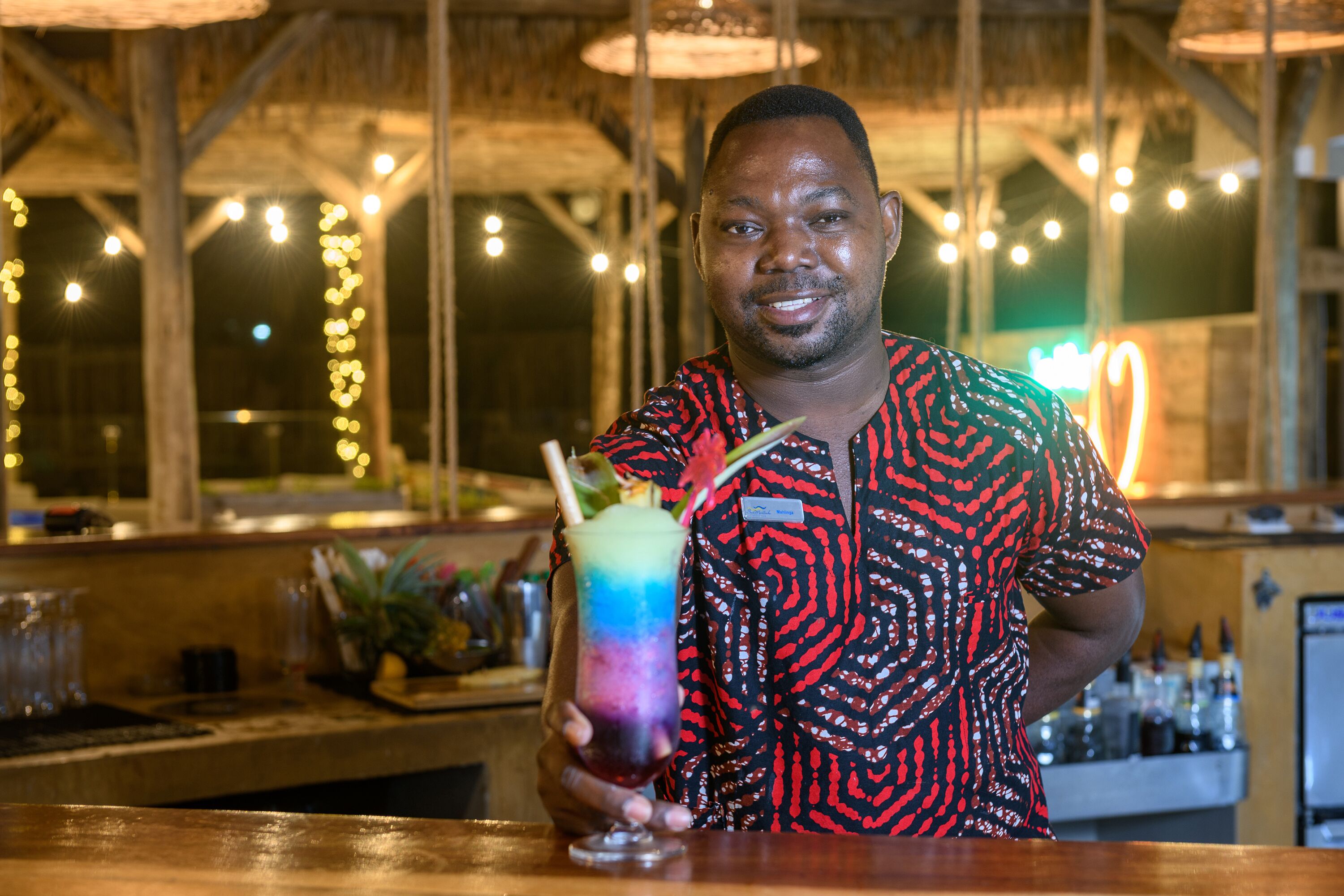 A person holding a colorful drink at a bar with wooden beams and string lights in the background.