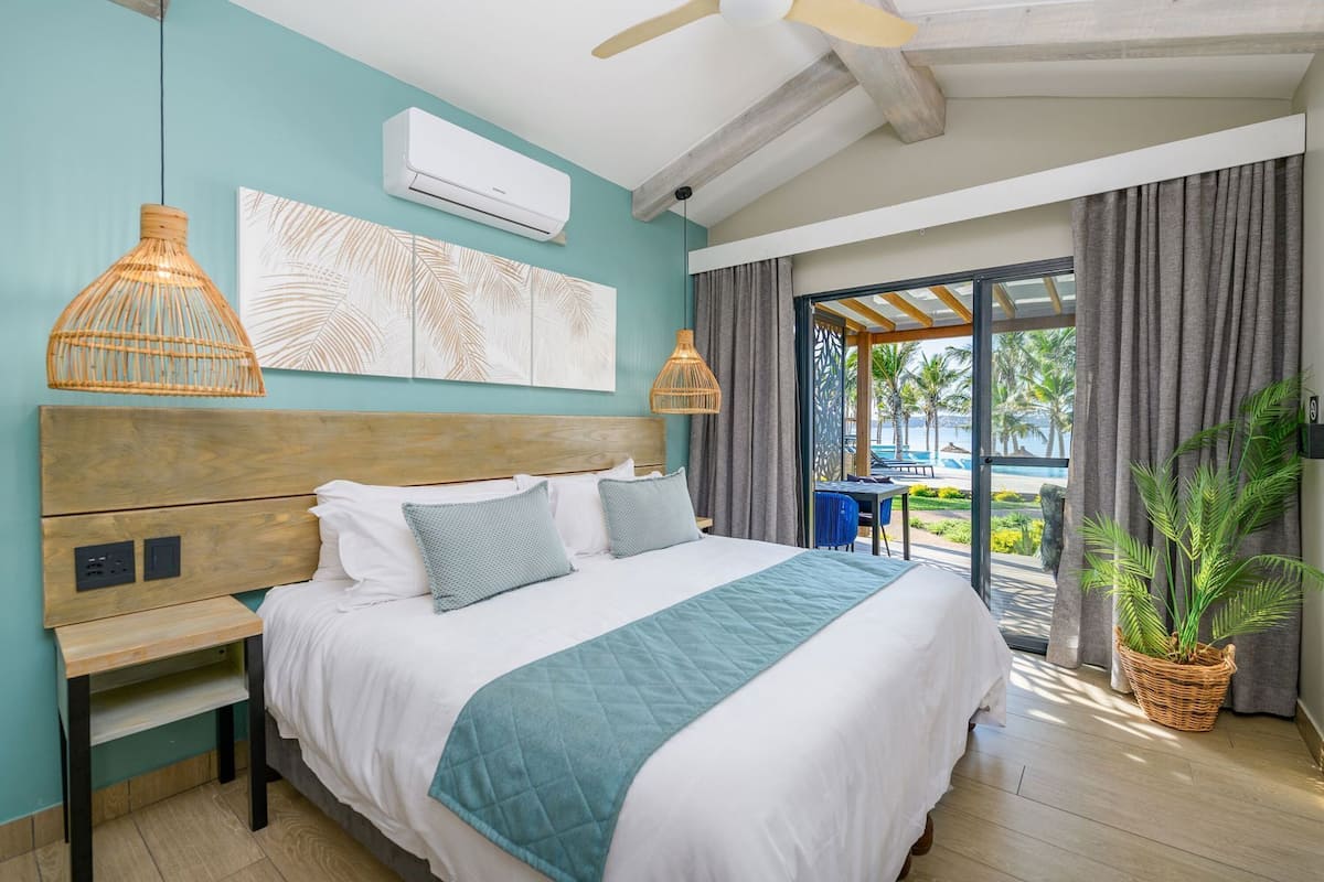 A hotel room with a bed, bedside table, wall-mounted air conditioner, ceiling fan, and a view of the pool and palm trees.