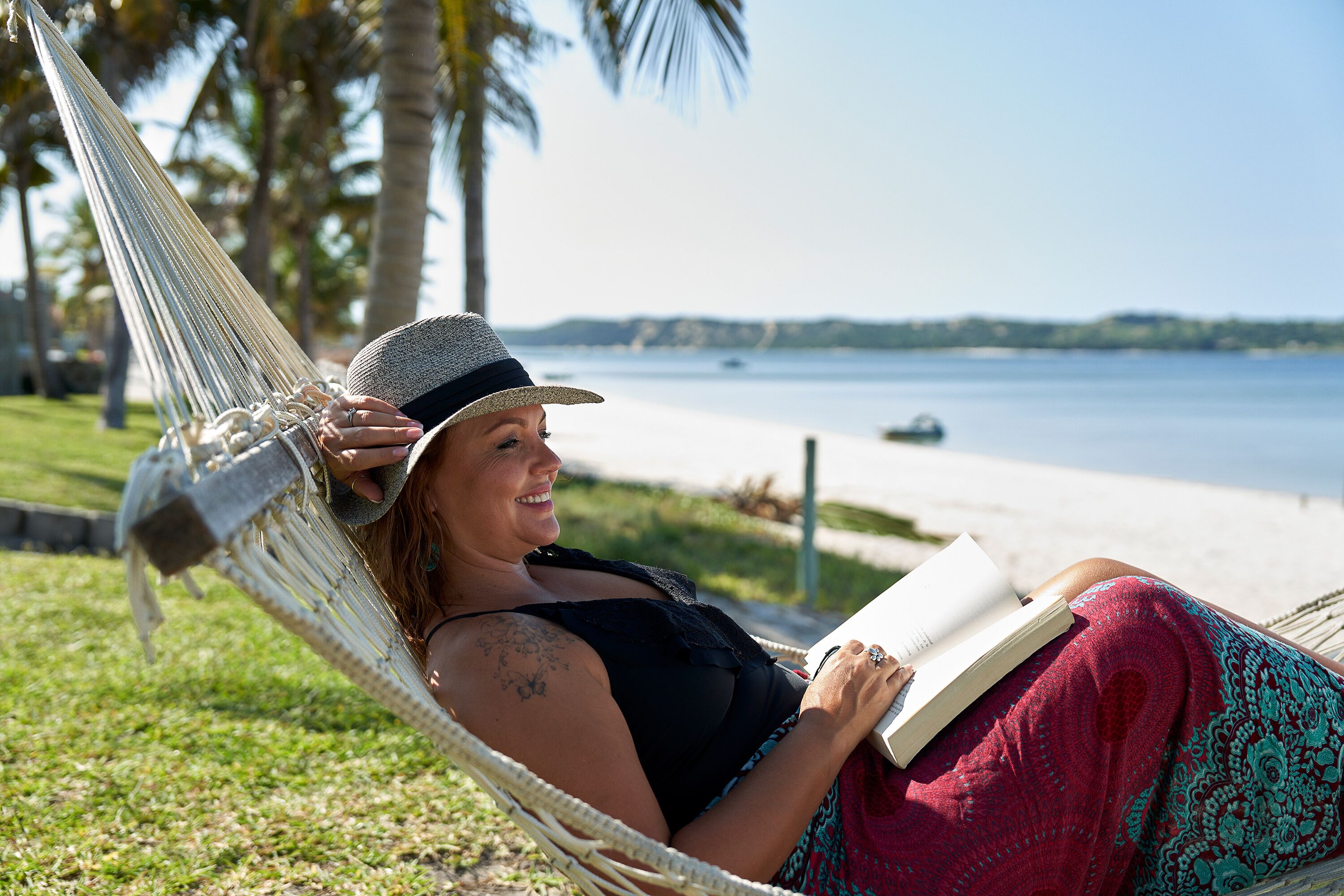 A person relaxing in a hammock on a grassy area with a beach and palm trees in the background.